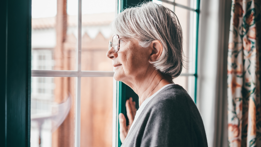 Alte ältere, grauhaarige Frau mit einer Brille, die am Fenster steht und nach draußen schaut. Senior Frau melancholische Träume zu Hause mit nachdenklichem Ausdruck.