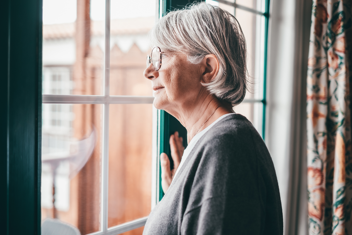 Alte ältere, grauhaarige Frau mit einer Brille, die am Fenster steht und nach draußen schaut. Senior Frau melancholische Träume zu Hause mit nachdenklichem Ausdruck.