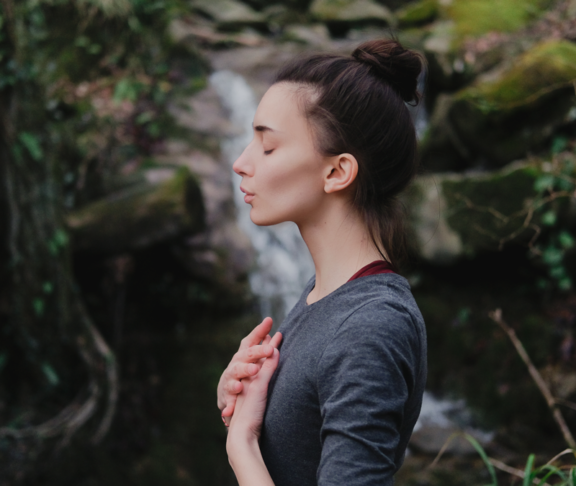Junge Frau, die im Mooswald auf dem Hintergrund des Wasserfalls Yoga pranayama draußen atmet. Einheit mit Naturkonzept