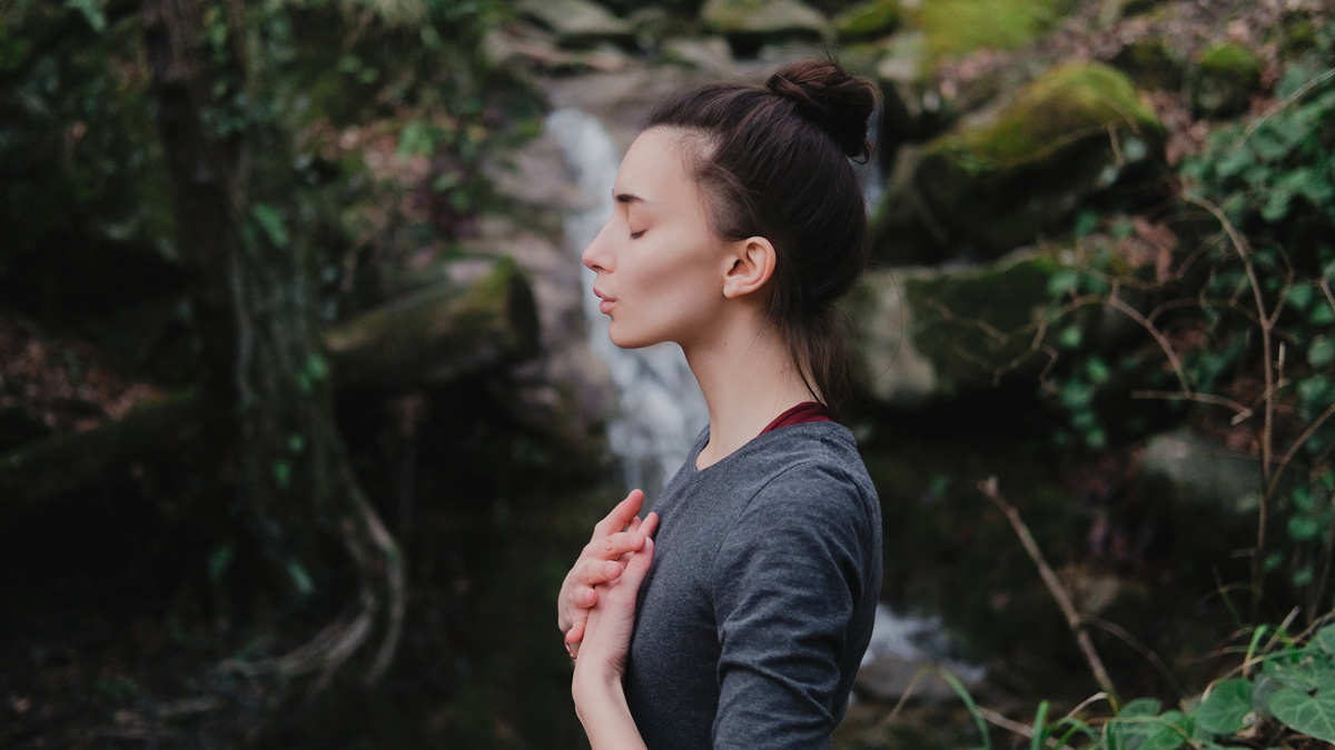 Junge Frau, die im Mooswald auf dem Hintergrund des Wasserfalls Yoga pranayama draußen atmet. Einheit mit Naturkonzept