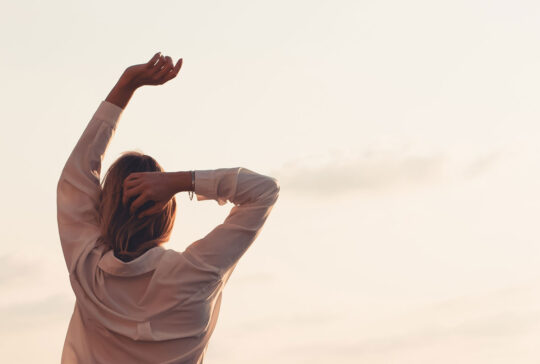 ​Rückansicht einer jungen Frau, die ihre Arme in Richtung eines sanften Sonnenuntergangshimmels streckt. Ein friedlicher und erstrebenswerter Lebensstil, der Freiheit, Wellness, Achtsamkeit und den Beginn eines neuen Tages oder eines ruhigen Abends repräsentiert.