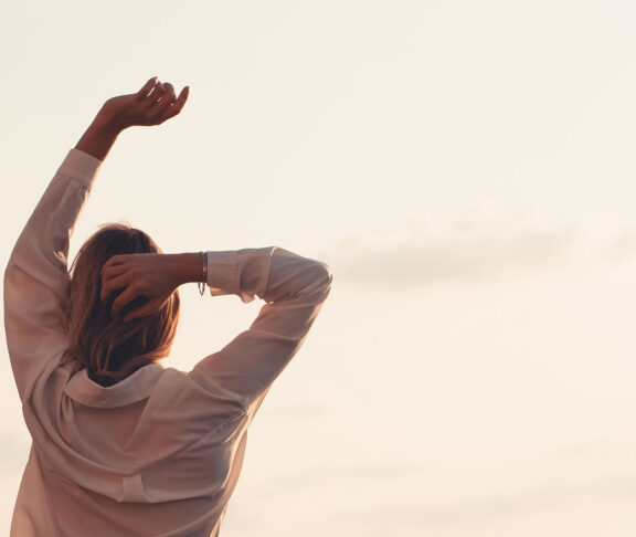 ​Rückansicht einer jungen Frau, die ihre Arme in Richtung eines sanften Sonnenuntergangshimmels streckt. Ein friedlicher und erstrebenswerter Lebensstil, der Freiheit, Wellness, Achtsamkeit und den Beginn eines neuen Tages oder eines ruhigen Abends repräsentiert.