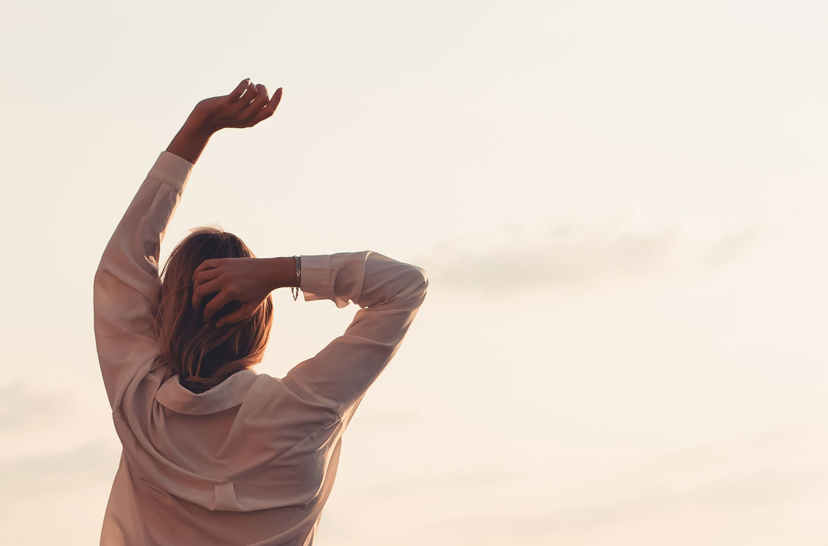 ​Rückansicht einer jungen Frau, die ihre Arme in Richtung eines sanften Sonnenuntergangshimmels streckt. Ein friedlicher und erstrebenswerter Lebensstil, der Freiheit, Wellness, Achtsamkeit und den Beginn eines neuen Tages oder eines ruhigen Abends repräsentiert.