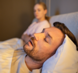 Mann schlafend im Bett und schnarchen laut, während frustrierte Frau neben sitzen mit erschöpften Ausdruck, gestresste Frau verliert Schlaf wegen Ehemann schnarcht in der Nacht, Gesundheit und Beziehungen Probleme.