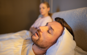 Mann schlafend im Bett und schnarchen laut, während frustrierte Frau neben sitzen mit erschöpften Ausdruck, gestresste Frau verliert Schlaf wegen Ehemann schnarcht in der Nacht, Gesundheit und Beziehungen Probleme.
