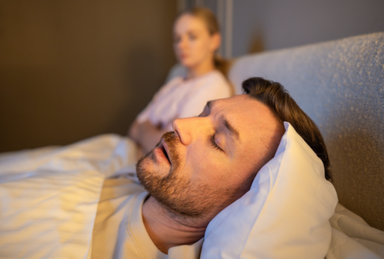 Mann schlafend im Bett und schnarchen laut, während frustrierte Frau neben sitzen mit erschöpften Ausdruck, gestresste Frau verliert Schlaf wegen Ehemann schnarcht in der Nacht, Gesundheit und Beziehungen Probleme.