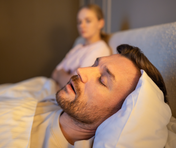 Mann schlafend im Bett und schnarchen laut, während frustrierte Frau neben sitzen mit erschöpften Ausdruck, gestresste Frau verliert Schlaf wegen Ehemann schnarcht in der Nacht, Gesundheit und Beziehungen Probleme.