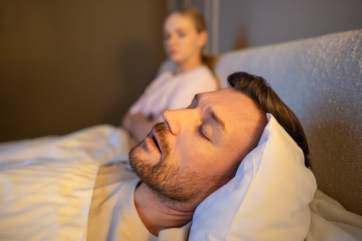 Mann schlafend im Bett und schnarchen laut, während frustrierte Frau neben sitzen mit erschöpften Ausdruck, gestresste Frau verliert Schlaf wegen Ehemann schnarcht in der Nacht, Gesundheit und Beziehungen Probleme.