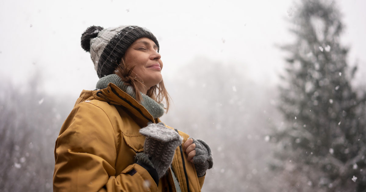 Young mature adult caucasian woman in a hat and yellow jacket breathing fresh air in the winter forest