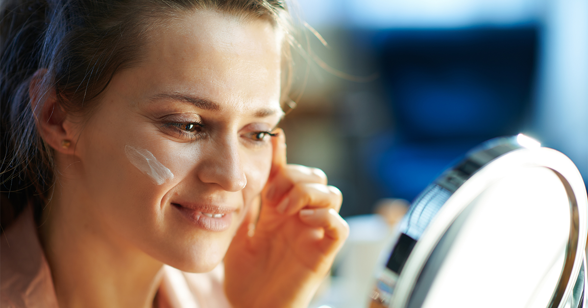 Elegant female applying facial cream and looking in mirror