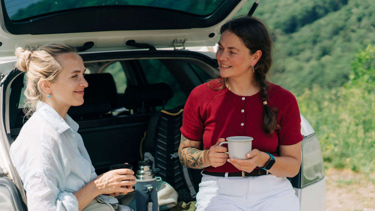 Two women friends are sitting in the back of car, talking and drinking coffee from a thermos during a trip out of city