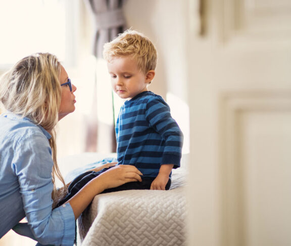 A young mother talking to her toddler son inside in a bedroom.