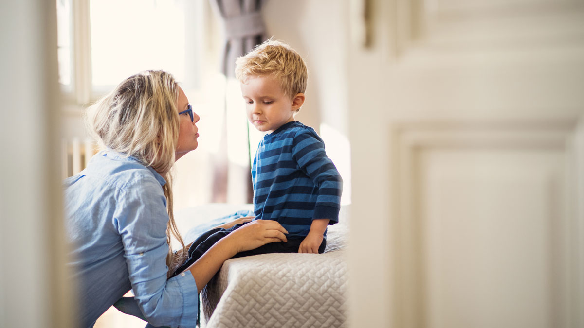 A young mother talking to her toddler son inside in a bedroom.