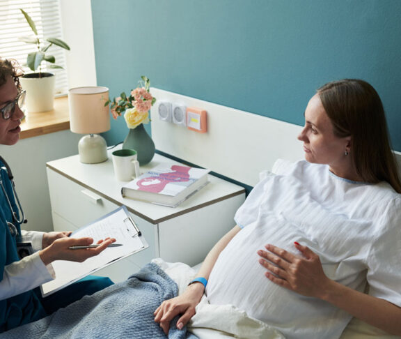 Caucasian pregnant young adult woman lying in hospital bed talking with Caucasian middle aged female doctor holding clipboard, medical consultation during pregnancy checkup