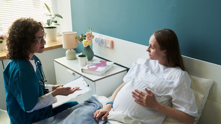 Caucasian pregnant young adult woman lying in hospital bed talking with Caucasian middle aged female doctor holding clipboard, medical consultation during pregnancy checkup