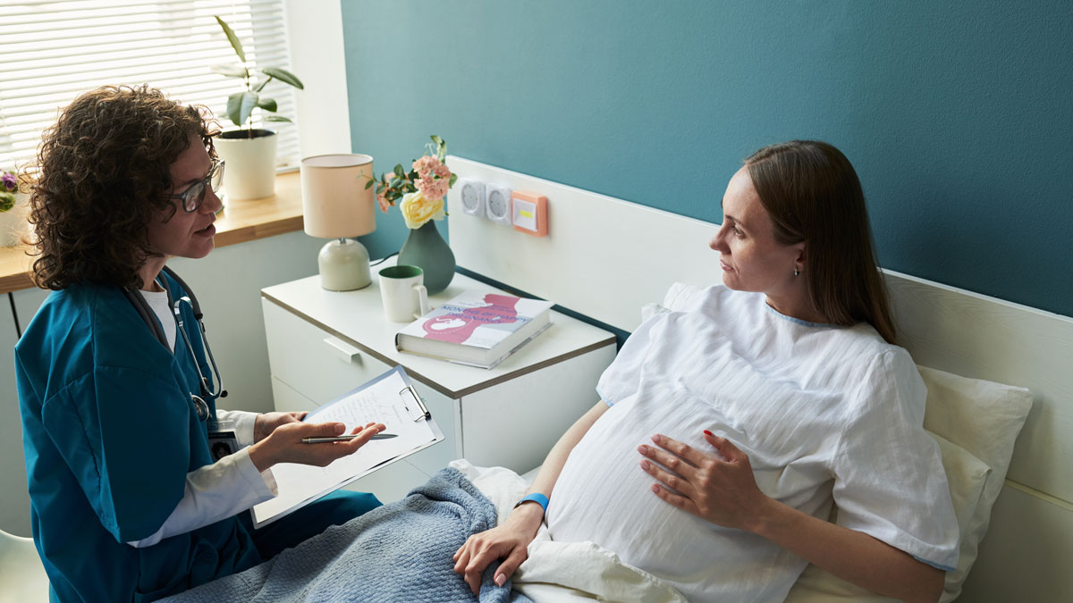 Caucasian pregnant young adult woman lying in hospital bed talking with Caucasian middle aged female doctor holding clipboard, medical consultation during pregnancy checkup