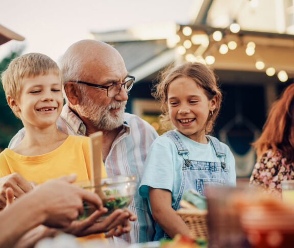 Happy Senior Grandfather Talking and Having Fun with His Grandchildren, Holding Them on Lap at a Outdoors Dinner with Food and Drinks. Adults at a Garden Party Together with Kids