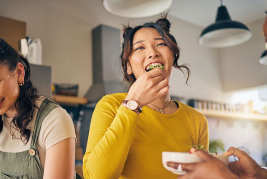 Women, friends and eating healthy food in home for bonding, nutrition and happy lunch together. Fruit diet, sharing and wellness, fresh summer friendship and girls in kitchen with smile at party.