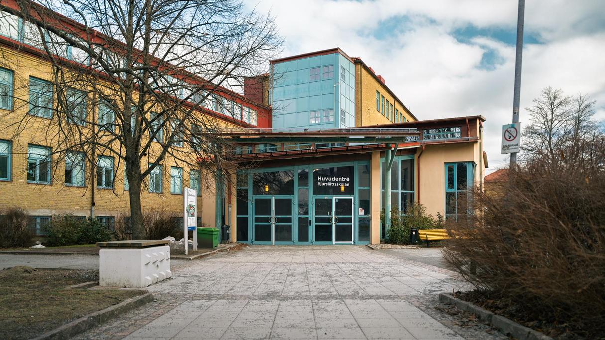 En entré till en skolbyggnad med texten Huvudentré Bjurslättsskolan på glasdörren och en gångväg som leder till den.