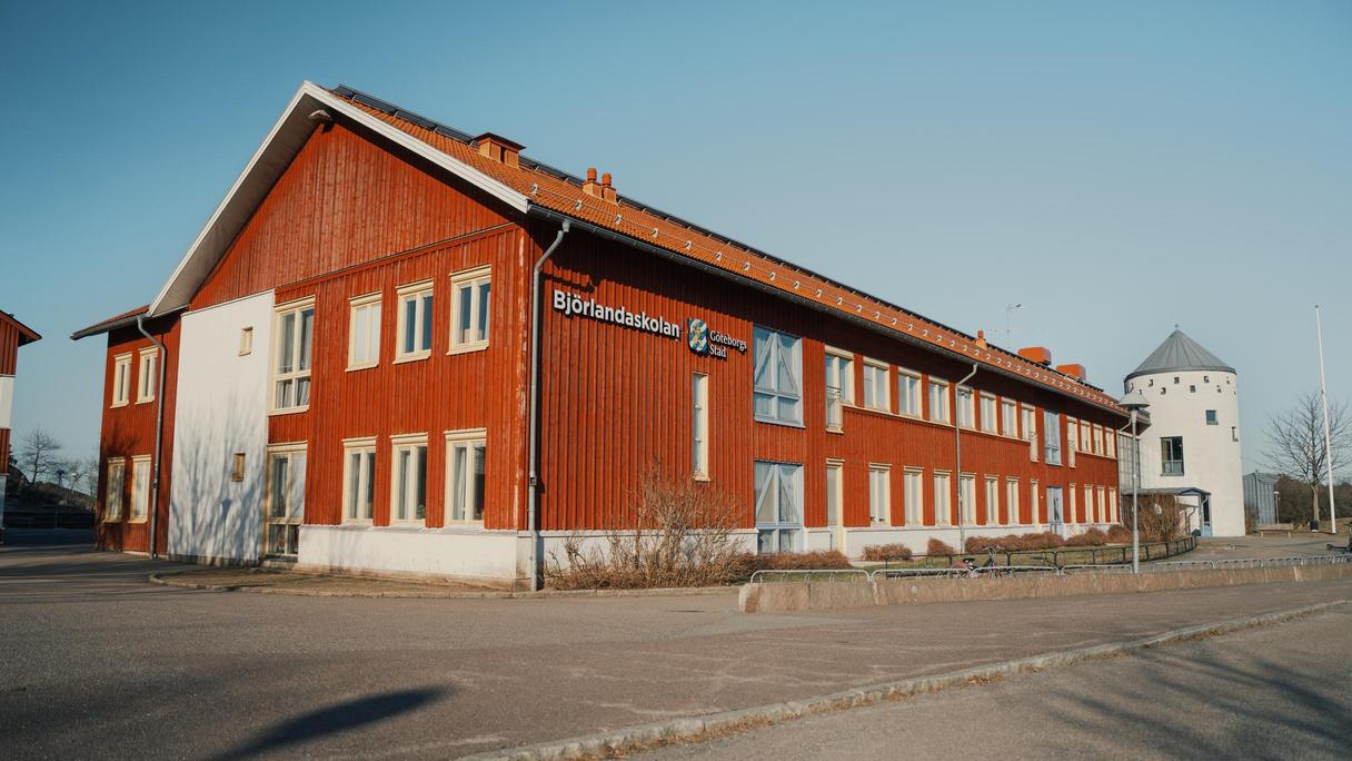 En stor röd skolbyggnad i trä med texten Björlandaskolan på fasaden som står vid sidan av en väg.