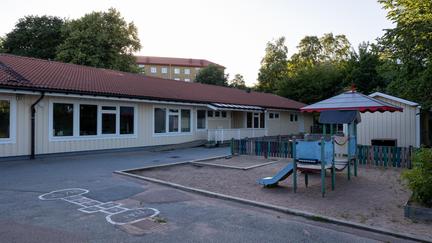 Bilden föreställer en ljusgul byggnad med vita fönsterkarmar. Framför byggnaden finns et förråd och en klätterställning med ruschkana.