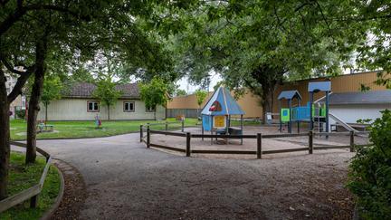 Bilden föreställer en lekpark med en blå rutschkana och ett blått lekhus. Runt parken finns ett brunt staket. Bakom lekparken finns ett grå-beige hus med röda fönsterkarmar.