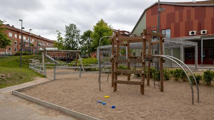 Bilden föreställer en lekplats med en klätterställning i trä och en kompisgunga. Bakom lekplatsen finns en förskola med röd och grön husfasad,