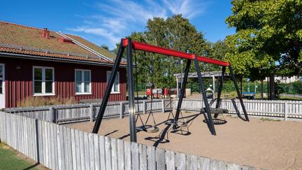 Bilden föreställer en röd träbyggnad med vita fönsterkarmar. Framför byggnaden finns en lekplats med tre gungor. Längre bak i bild syns n klätterställning med ruschkana.