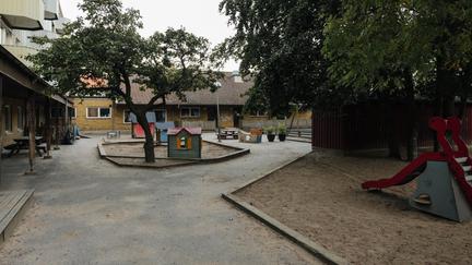 Bilden föreställer en tegelbyggnad. Framför byggnaden finns lekplats med tre leksaksstugor, en ruschkana, utomhusbänkar och gungställning.