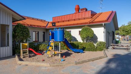 Bilden föreställer en vit byggnad och en stor blå och en mindre röd ruschkana i en sandlåda.