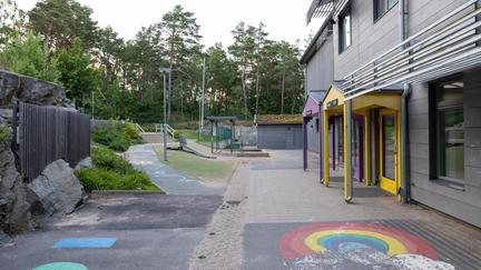 Bilden föreställer en grå byggnad med en lila och en gul dörr Framför byggnaden finns en sandlåda och en ruschkana. En regnbåge är ritad på asfalten.