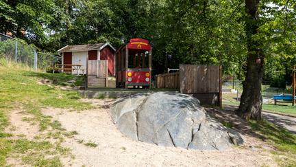 Bilden föreställer en lekplats på gräs. Bredvid ett träd finns en stor sten och bakom stenen finns en stor röd leksaksbil och en röd stuga. 