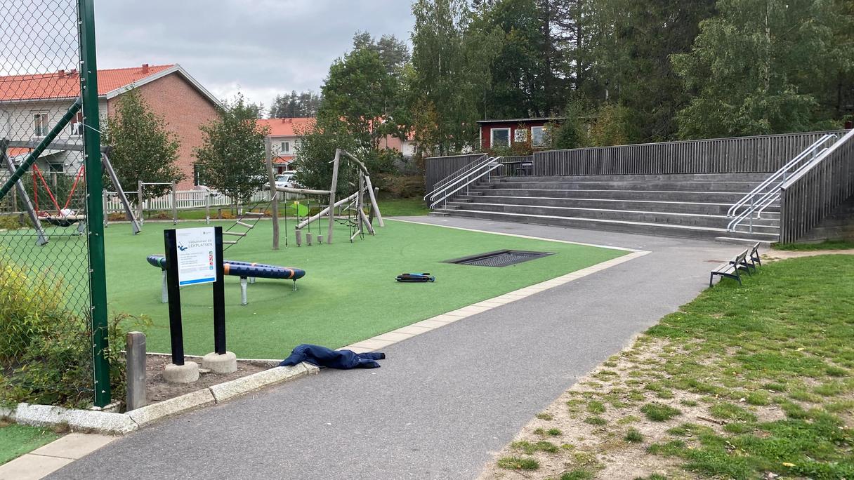 Öppen lekplats med många lekredskap på gummiasfalt. Stor läktare i trä och skogsdunge bakom lekplatsen