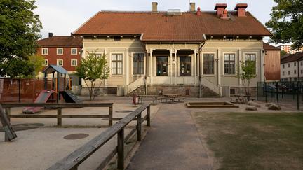 Bilden föreställer ett trähus med ett tegeltak. Framför huset finns en lekplats med en sandlåda, rutschkana, gräsmatta och staket.  