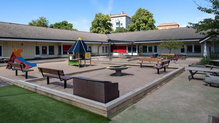 Bilden föreställer en ljus hörnbyggnad med rosa persienner. Framför byggnaden finns en lekplats med en lekstuga, en ruschkana, utomhusbänkar och gunghästar. 