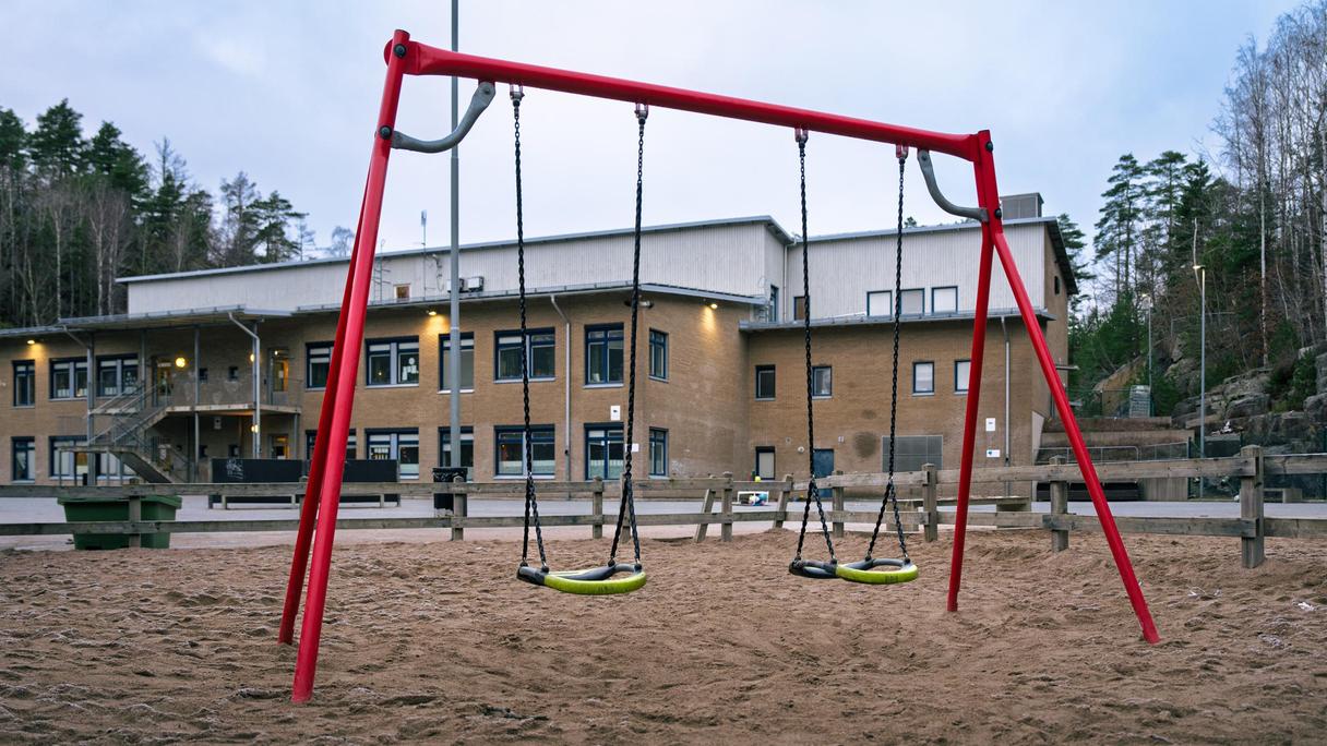 En röd gungställning i en sandlåda framför en skolbyggnad i gult tegel.