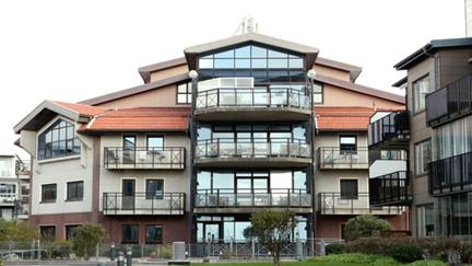 Ett modernt flerbostadshus med flera balkonger, stora glaspartier och rött tegeltak. Framför huset finns en gångväg, gräsmatta och buskar.