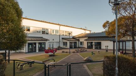 Bilden föreställer en vit byggnad med tre entréer. Framför byggnaden finns en inhägnad park med en leksaksbåt, gunghästar och parkbänk.
