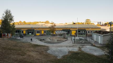 Bilden föreställer en gul byggnad med en förskolegård. På gården finns gungor, en sandlåda, förråd och träd.