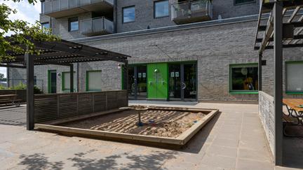 Bilden föreställer en grå tegelbyggnad med en grön dörr. Framför ingången finns en sandlåda med ett bord. 