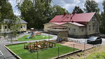 Bilden föreställer en gul byggnad med en förskolegård med en sandlåda, en ruschkana och krukor med växter.