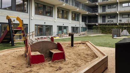 Bilden föreställer en sandlåda med en leksaksbil och en ruschkana. Bakom lekplatsen finns en grå byggnad.