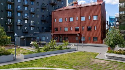 Bilden föreställer en röd byggnad och en grå byggnad. Framför byggnaderna finns en uteplats med träd och förråd.