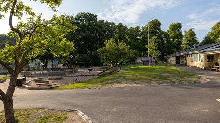 Bilden föreställer en gul träbyggnad. Framför byggnaden finns en lekplats med en gunga, klätterställning och rutschbana. Runt lekplatsen finns gröna träd.