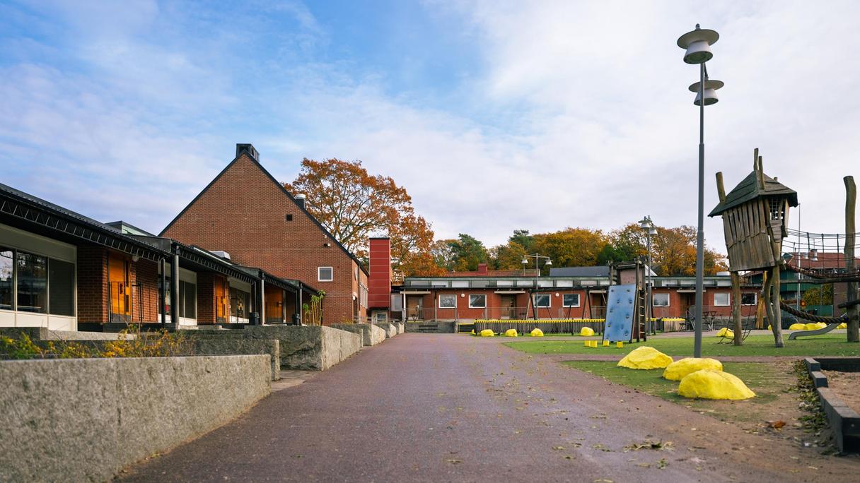 En väg som leder in mot Hovåsskolan ligger i fokus på bilden och skolgården i bakgrunden. Några gulmålade stenar och en klätterställning syns även i bakgrunden.