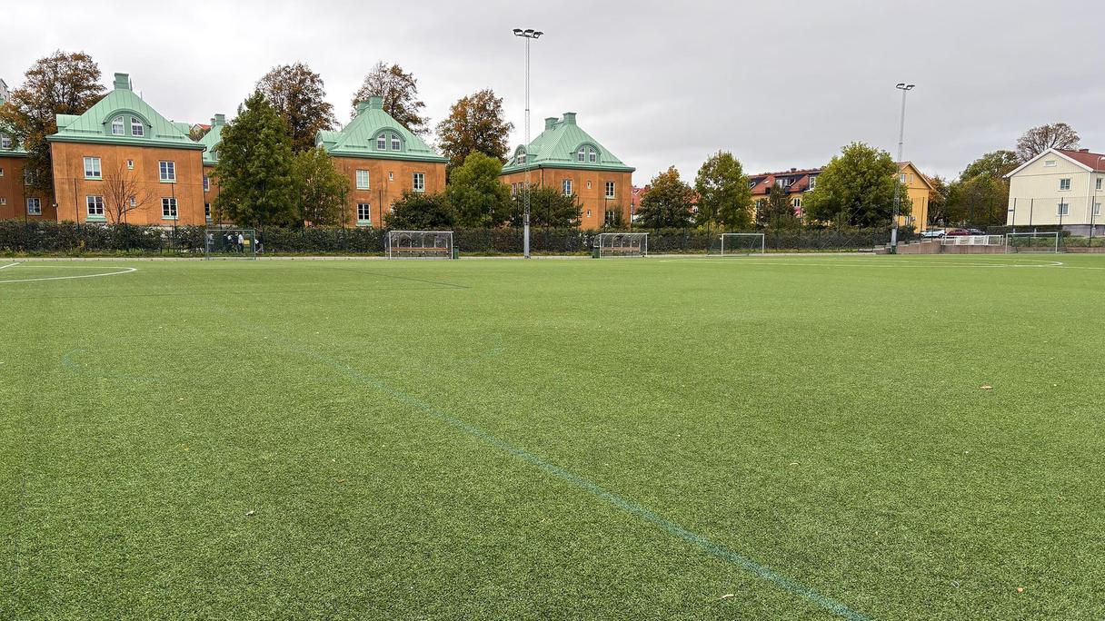 En grön fotbollsplan med Härlanda Parks byggnader i bakgrunden.