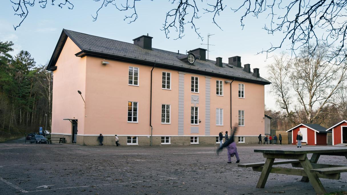 Huvudbyggnaden för Högenskolan med en parkbänk och några barn i bakgrunden.