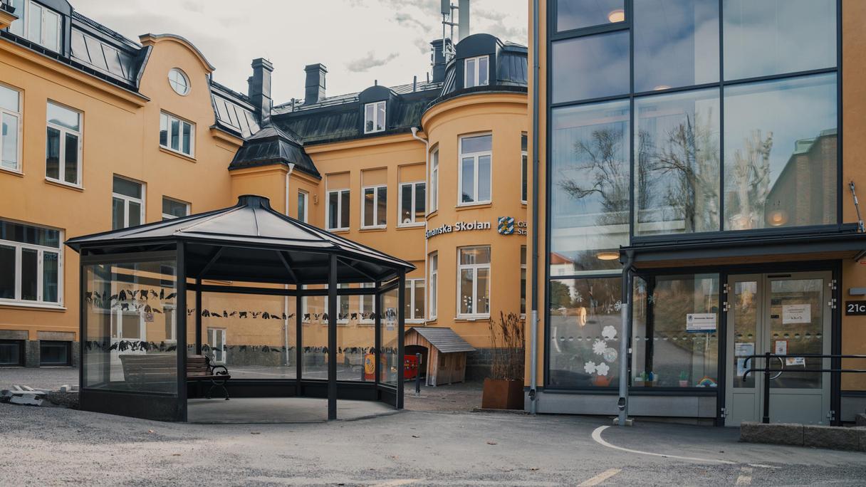 En paviljong med en bänk i framför en gul skolbyggnad med en entré