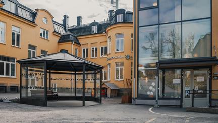 En paviljong med en bänk i framför en gul skolbyggnad med en entré