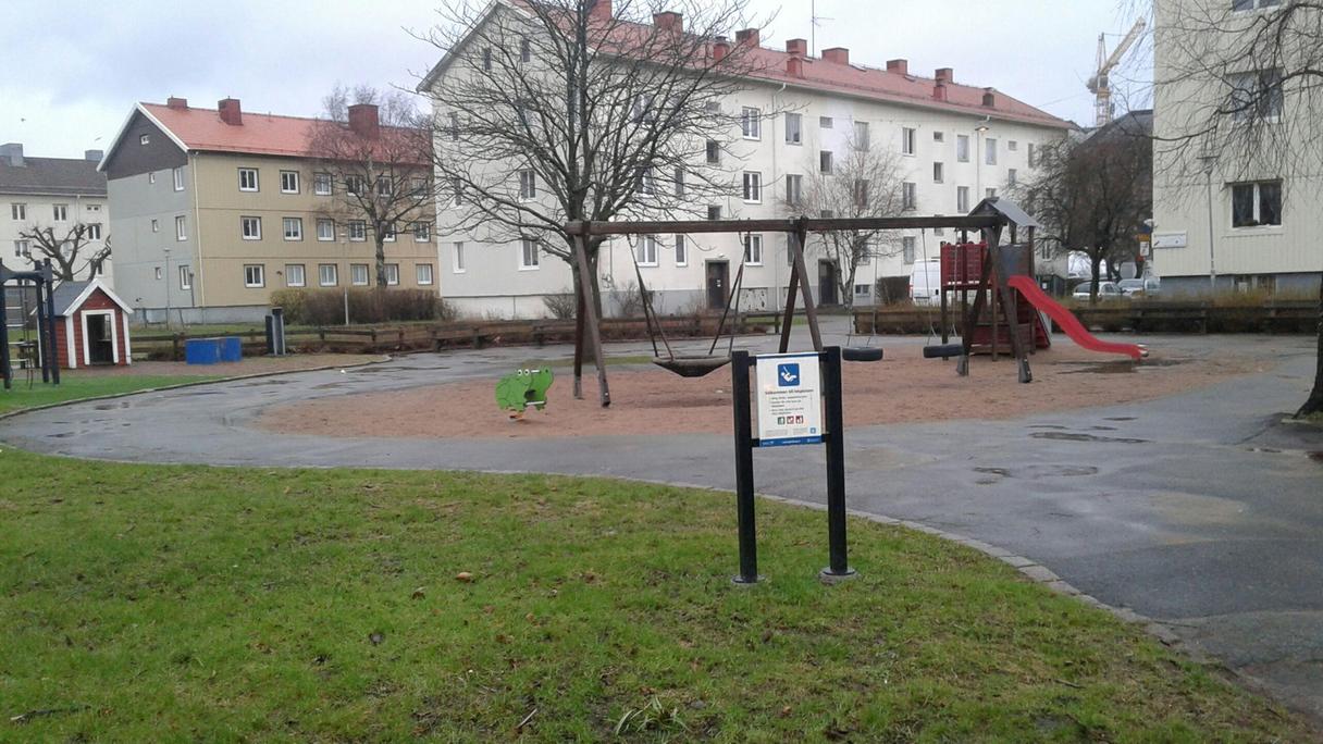 Lekplatsen ligger i ett bostadsområde, gräsmattor, asfalterad gångväg, bänkar och sandlåda med gungor och rutschkana